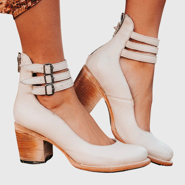 A woman wearing tan block-heel court shoes with buckled ankle straps, walking gracefully in a refined outfit.