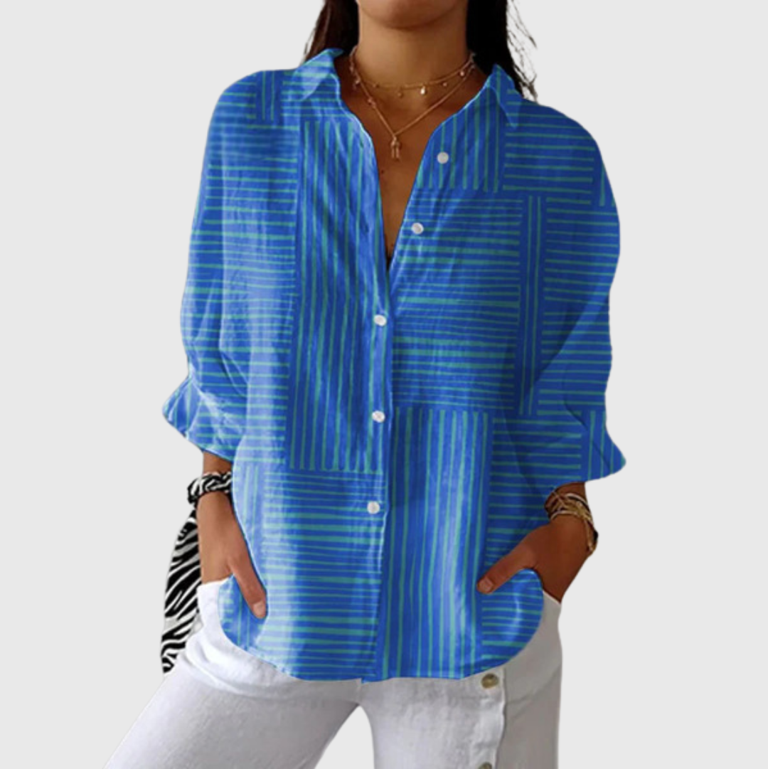A woman wearing a loose blue striped blouse with white trousers, relaxed and smiling