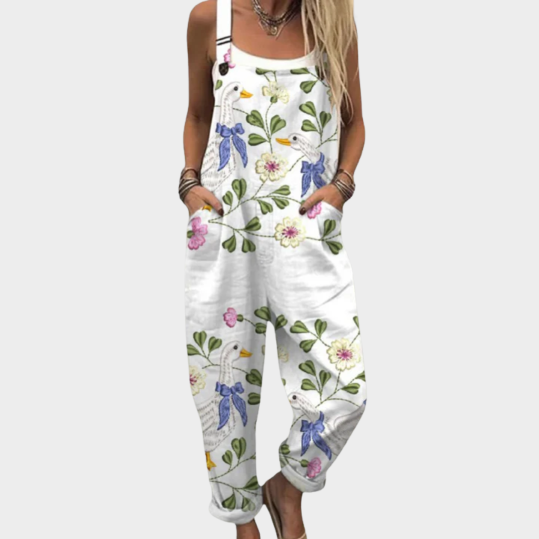 Woman wearing white dungarees printed with geese in blue bows among green leaves and blooms, adjustable straps, cheerful and relaxed.