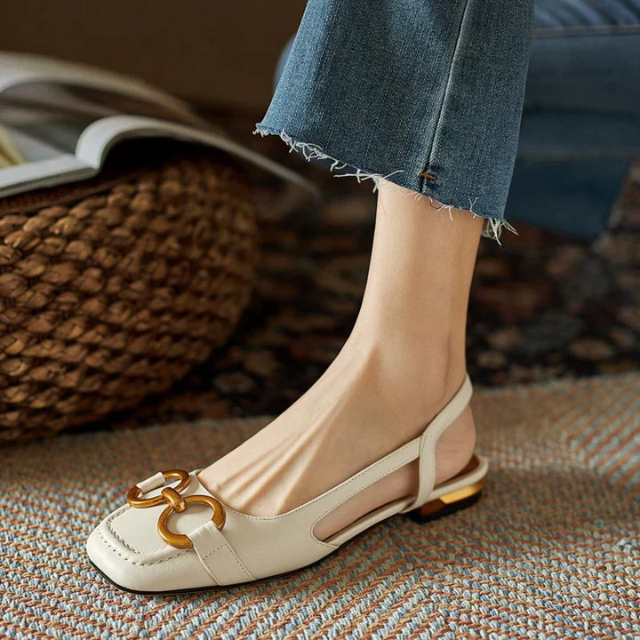 A woman wearing navy square-toe open-back flats with a small golden buckle detail.