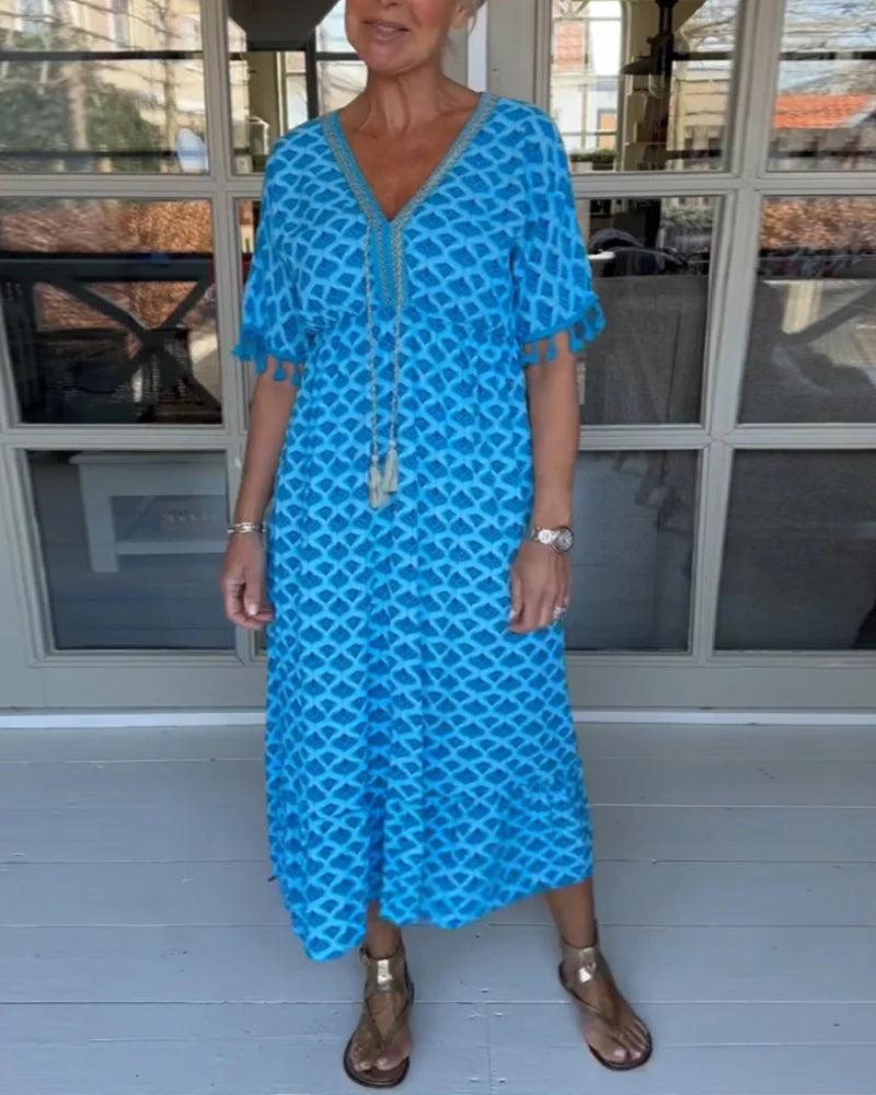 A woman walking near the coast in a flowing blue diamond-lattice print maxi dress with a tassel-trimmed neckline.