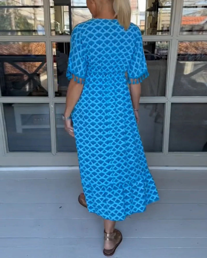 A woman walking near the coast in a flowing blue diamond-lattice print maxi dress with a tassel-trimmed neckline.