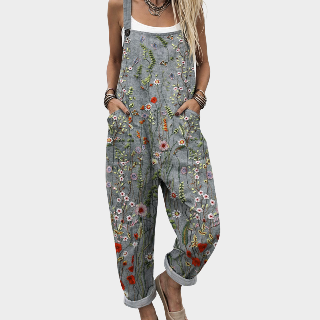 A woman wearing grey dungarees covered in bright meadow flowers, daisies and poppies, with adjustable straps.