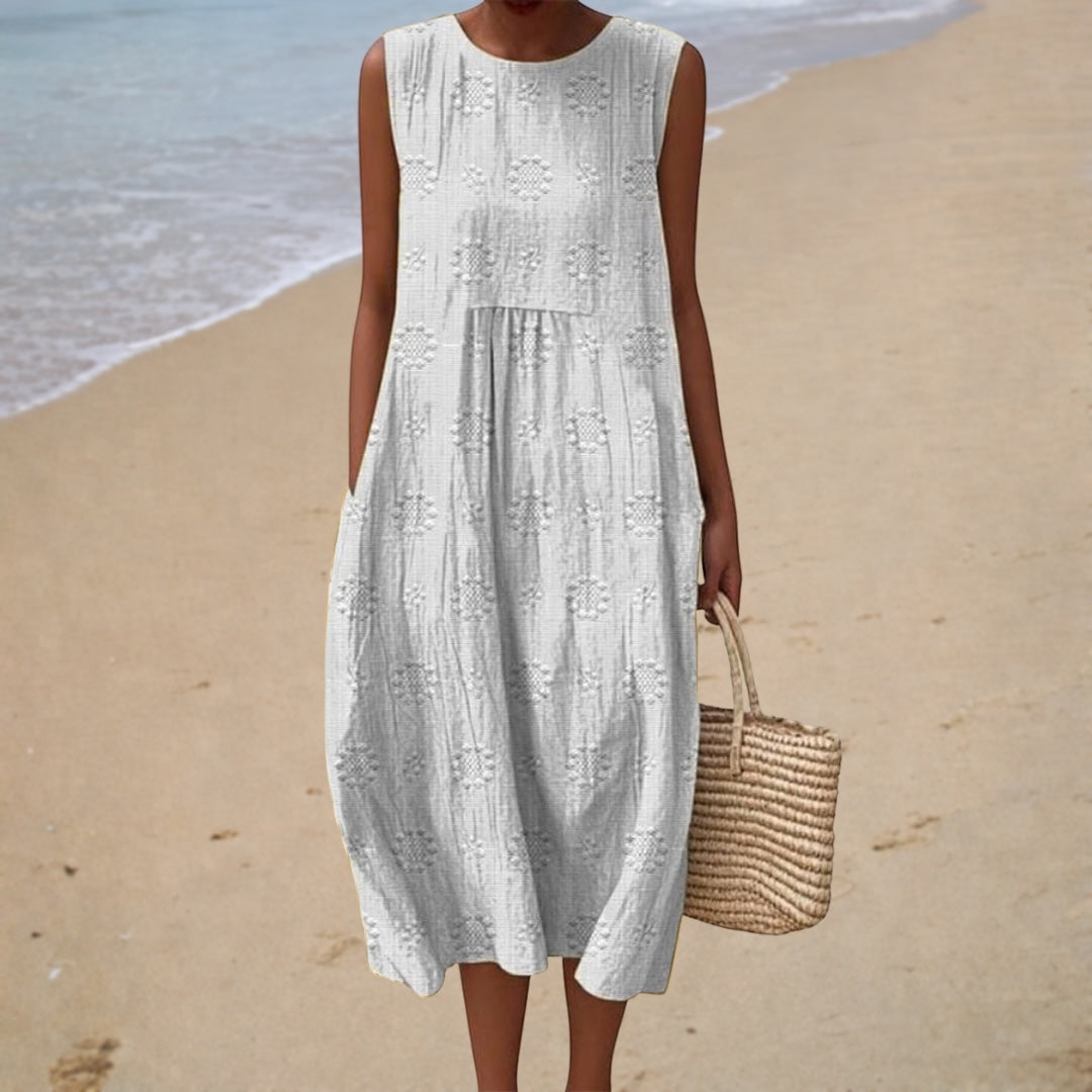 A woman on the beach wearing a soft white sleeveless summer dress with tonal woven dot detail.