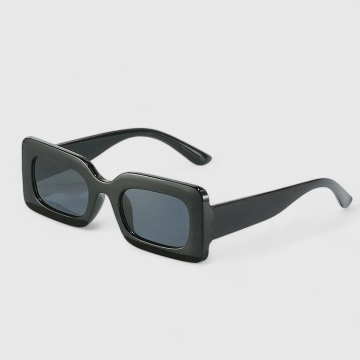 A woman on a sunny terrace wearing bold black rectangular sunglasses, poised and confident.