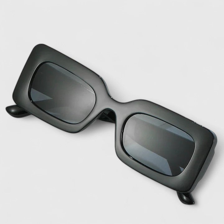 A woman on a sunny terrace wearing bold black rectangular sunglasses, poised and confident.