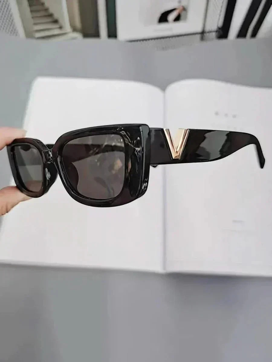 A woman at an outdoor event wearing chunky black rectangular retro sunglasses with a gold-tone accent.