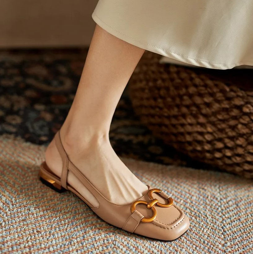 A woman wearing navy square-toe open-back flats with a small golden buckle detail.