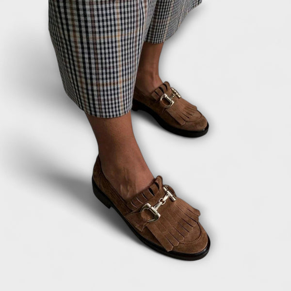 A woman wearing brown fringed loafers with a gold snaffle detail and checked trousers, looking polished and at ease.