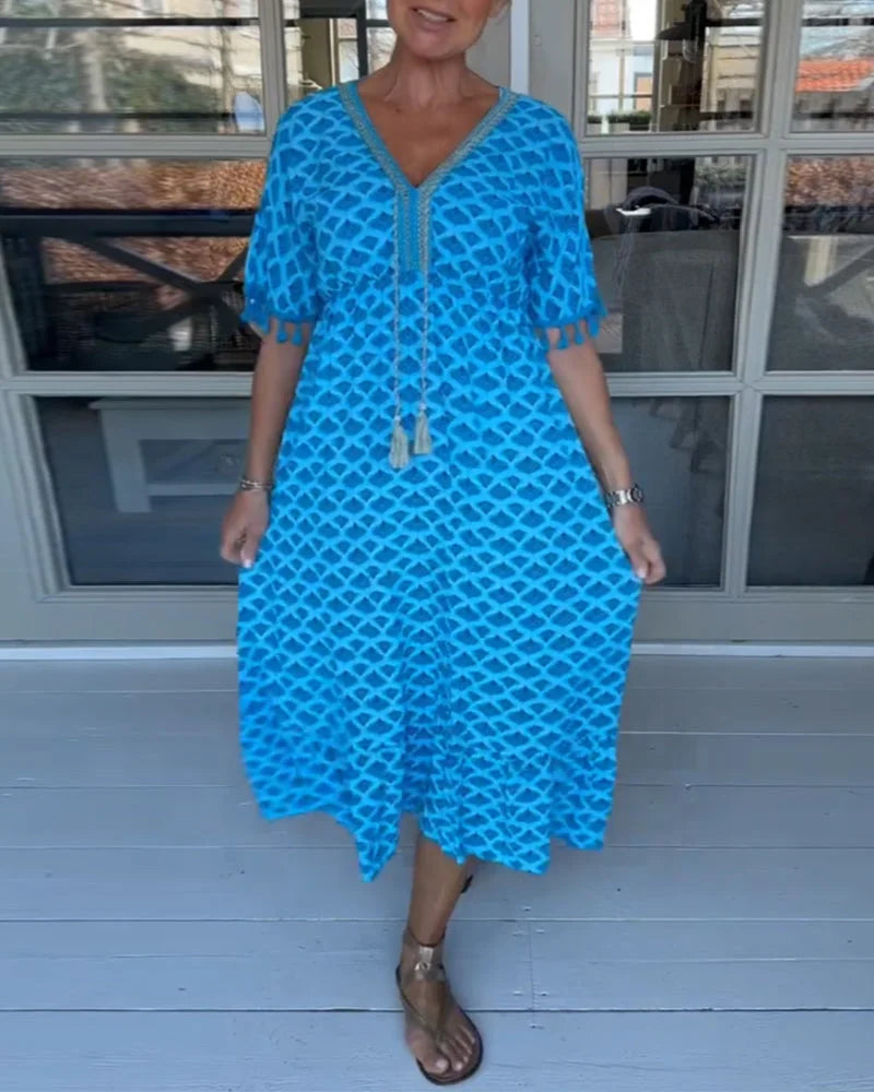 A woman walking near the coast in a flowing blue diamond-lattice print maxi dress with a tassel-trimmed neckline.