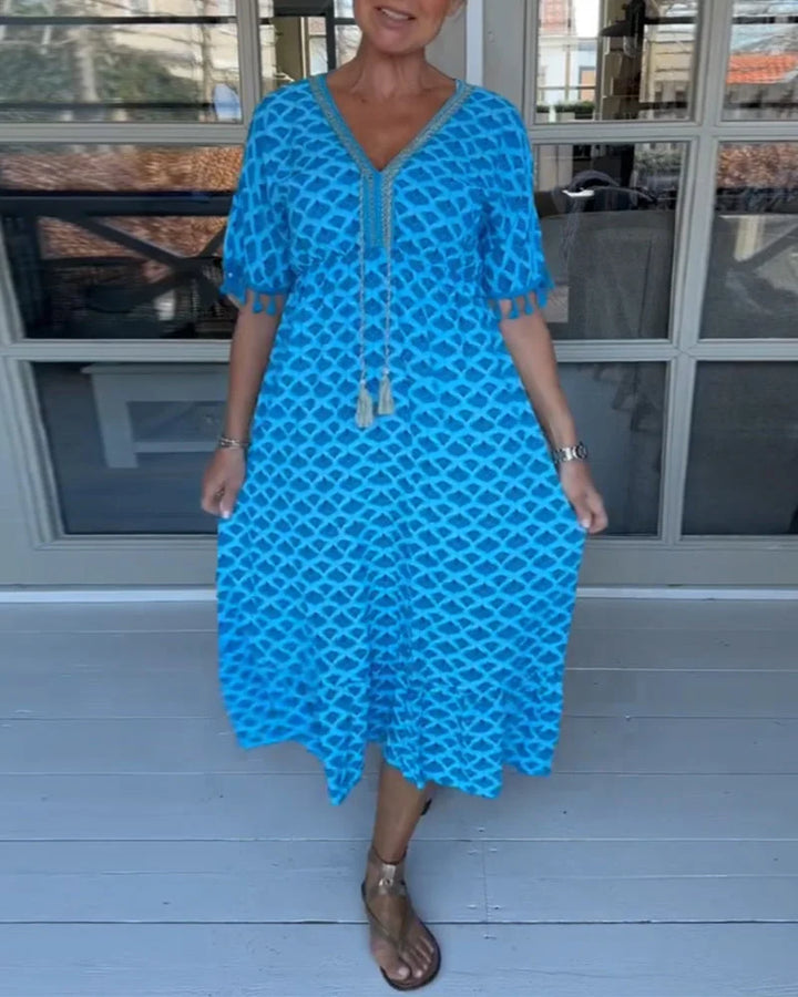 A woman walking near the coast in a flowing blue diamond-lattice print maxi dress with a tassel-trimmed neckline.