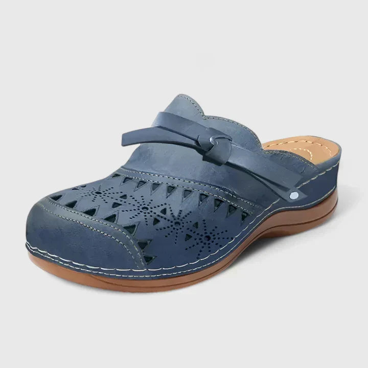 A woman slipping on airy laser-cut comfort shoes with a decorative knot detail at home.