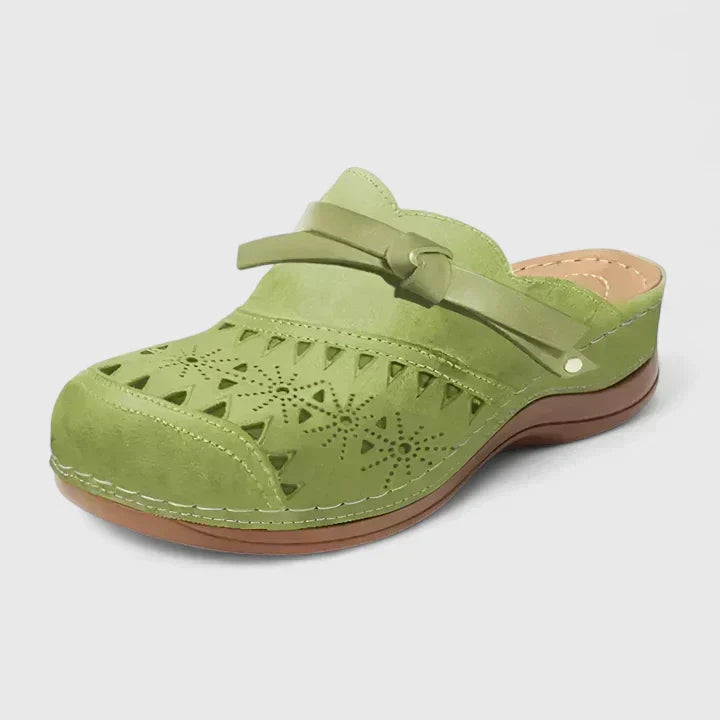 A woman slipping on airy laser-cut comfort shoes with a decorative knot detail at home.