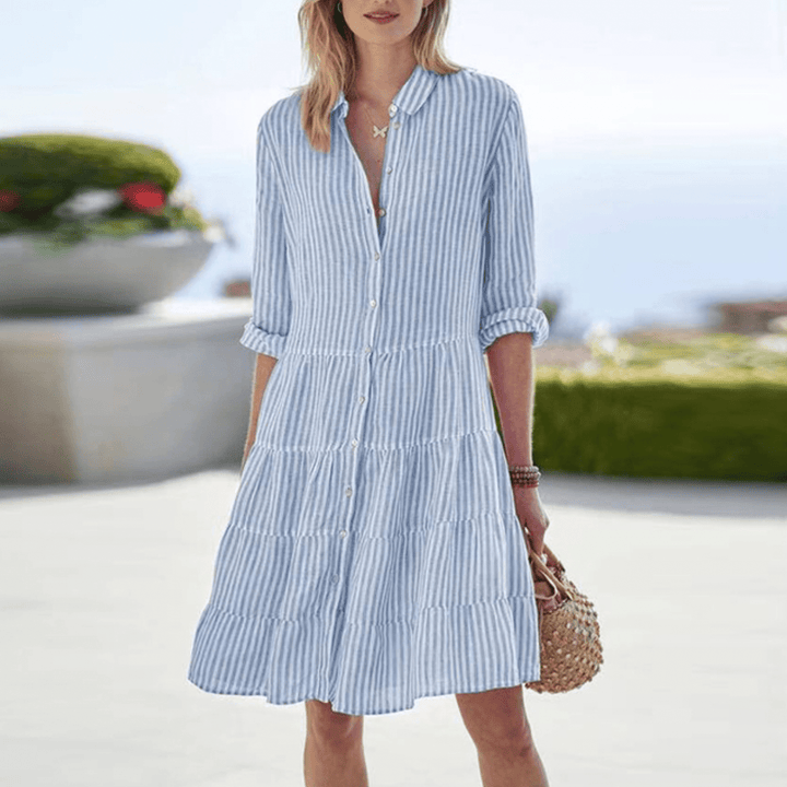 A woman wearing the Dympna coastal stripe dress with a tiered skirt and button front, strolling by the sea.